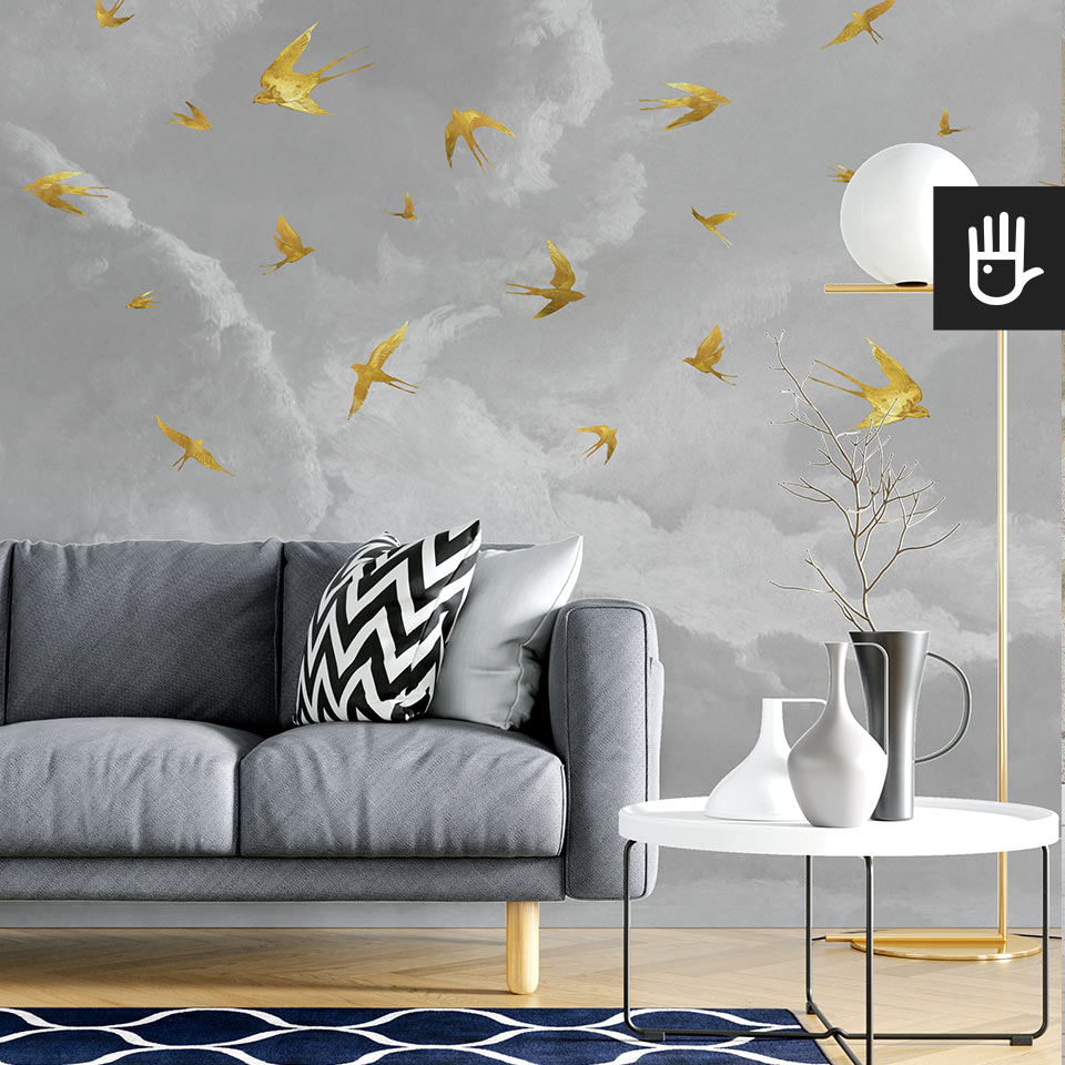 Nowoczesny salon w stylu skandynawskim z szarą kanapą na tle szarej ściany na której jest naklejona fototapeta Złote jaskółki ze złotymi ptakami na tle kłębiastych chmur.